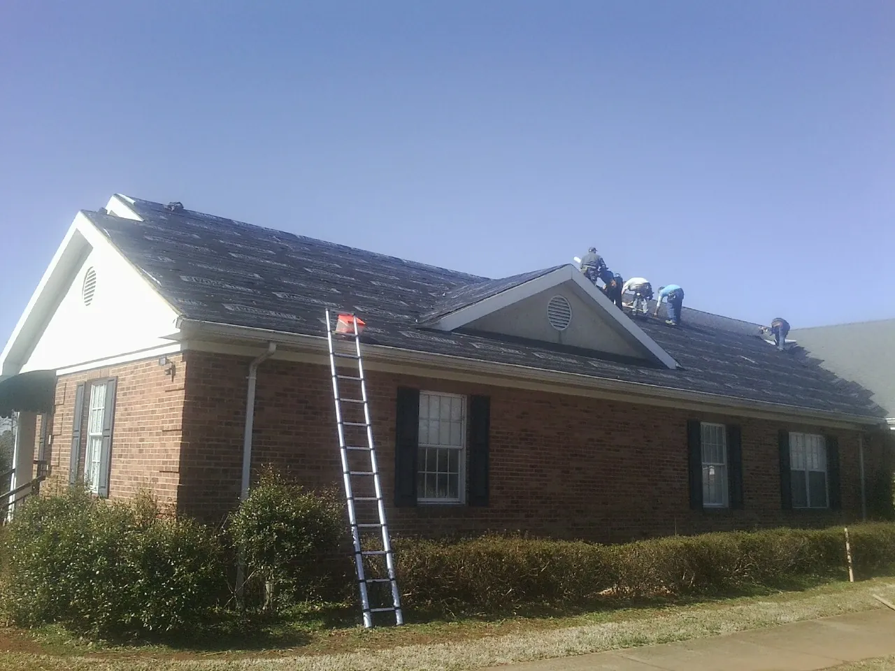 Crew tearing shingles off a brick ranch roof with a ladder set — Emergency Roof Repair work by our crew in Caledonia